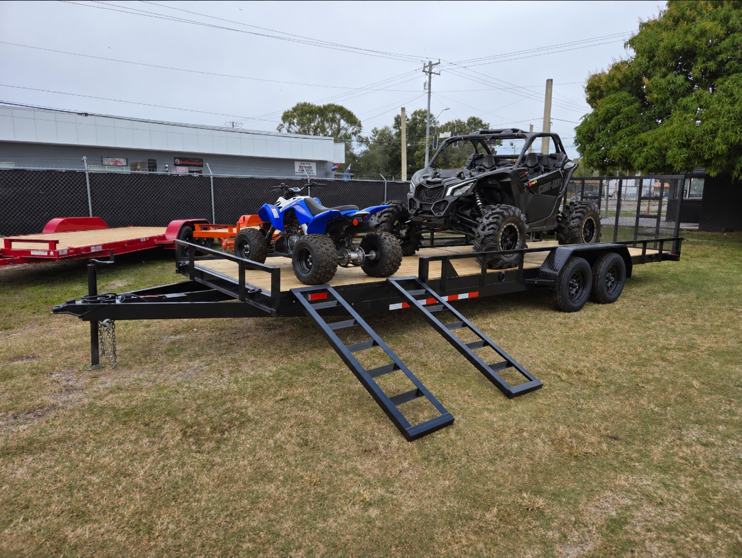 Utility trailer carrying a side-by-side and a smaller blue ATV above the side loading ramps